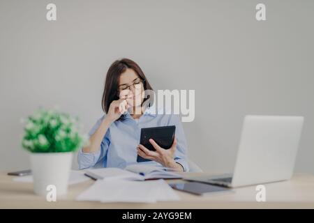 Cheerful female financial manager holds calculator in hands, being satisfied with recieved figures, makes report on laptop computer, analyzes company` Stock Photo